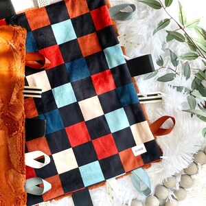 Puede incluir: Una manta de bebé a cuadros con cuadrados rojos, negros, blancos y azules. La manta tiene varios lazos y cintas de tela unidos a los bordes. La manta está hecha de tela suave y afelpada.