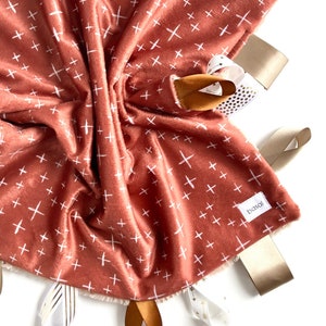 Puede incluir: Una manta sensorial para bebés de color terracota con signos más blancos y múltiples cintas de satén beige y doradas atadas. La manta tiene una etiqueta "bassi" en la esquina inferior derecha.