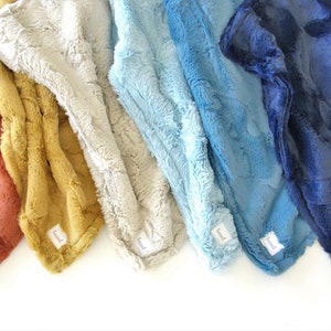May include: Five plush blankets in different colors, including rust, mustard yellow, beige, light blue, and navy blue. Each blanket has a small white tag with the word "boo" printed on it.