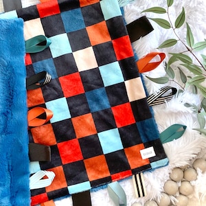 Puede incluir: Una manta para bebé a cuadros azules y blancos con etiquetas de tela de colores. La manta está hecha de tela suave y afelpada y tiene una variedad de texturas para que los bebés exploren. Las etiquetas están unidas a la manta con costuras y están hechas de diferentes materiales, incluyendo satén, terciopelo y algodón.