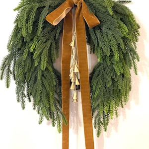 May include: A circular evergreen wreath with a brown velvet ribbon and small gold bells. The wreath is made of green foliage and has a full appearance. The ribbon is tied in a bow at the top, with long streamers.