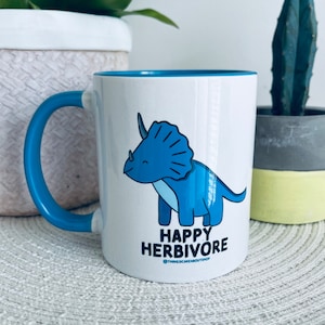 Puede incluir: Taza de cerámica blanca con borde y asa azules. La taza presenta un triceratops azul de dibujos animados con el texto "Happy Herbivore" y el nombre de usuario de las redes sociales "@thingsicareaboutshop".