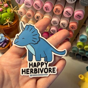 May include: A die-cut sticker featuring a light blue triceratops dinosaur with a dark blue underbelly and a smiling expression. Below the dinosaur, the text reads HAPPY HERBIVORE and @THINGSICAREABOUTSHOP in bold black capital letters.
