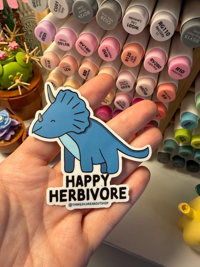 May include: A die-cut sticker featuring a light blue triceratops dinosaur with a dark blue underbelly and a smiling expression. Below the dinosaur, the text reads HAPPY HERBIVORE and @THINGSICAREABOUTSHOP in bold black capital letters.
