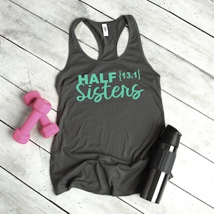 May include: Gray tank top with the text "HALF {13.1} Sisters" in teal green lettering. The tank top is laying on a white wood surface with pink dumbbells and a black water bottle.