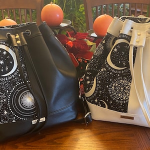 May include: Two black and white drawstring bucket bags with a crescent moon and star pattern. The bags have black leather accents and adjustable straps.
