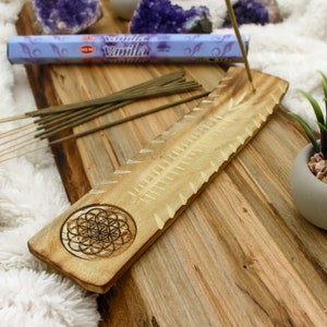 May include: A wooden incense holder with a carved geometric design and a series of angled notches. A box of "Vanilla" incense sticks and several brown incense sticks are placed on a wooden surface. Amethyst crystals and a small potted plant are in the background.