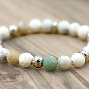 May include: A bracelet made of various natural stone beads in white, tan, and green, with silver-coloured metal beads. The bracelet is elasticated and displayed on a wooden surface. The beads are approximately 1 cm in diameter.