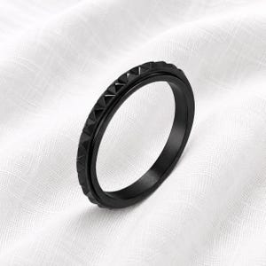 May include: A matte black ring featuring a textured outer band with a repeating pyramid stud pattern. The ring has a smooth inner surface and a consistent dark finish throughout, showcasing a geometric design with sharp, defined edges.