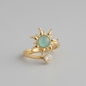 May include: A gold-toned open ring featuring a central sun-shaped motif with a smooth, round, pale mint-green cabochon stone. The opposite end of the adjustable band is finished with a single, small, clear, round-cut sparkling gemstone.
