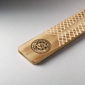 May include: A wooden incense burner with a carved design of a sun with a symbol inside. The burner has a grooved surface for burning incense.