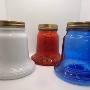May include: Three bell-shaped glass jars with metallic gold-toned lids. One jar is opaque white, one is translucent red, and one is translucent blue. Each jar features the embossed script text Kerr Self Sealing on the front surface.