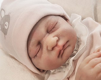 May include: A lifelike baby doll, dressed in a pink outfit with heart patterns, and a matching hat with a mouse design. The doll is lying on a soft, white blanket, with its eyes closed. The doll's skin has realistic details.