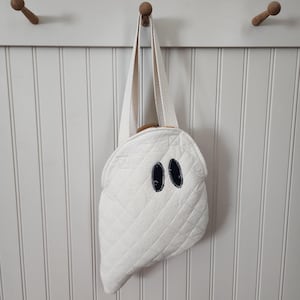 Pode incluir: Uma sacola tote branca acolchoada em forma de fantasma, com uma alça creme e olhos ovais azuis escuros. A sacola está pendurada em um pino de madeira em uma parede branca. A sacola tem um padrão acolchoado.