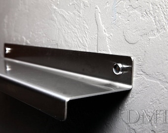 May include: A stainless steel shelf mounted on a textured gray wall. The shelf has a simple, modern design with a lip on the front edge. The shelf is attached to the wall with two screws.