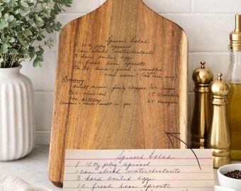 Pode incluir: Uma tábua de corte de madeira com uma alça, com notas de receitas manuscritas. Um cartão de receita com o título "Salada de Espinafre" está na frente da tábua. A tábua é marrom clara com grão de madeira natural.