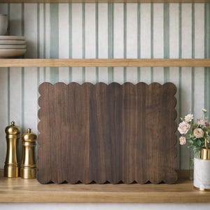 May include: A dark brown wooden cutting board with a scalloped edge sits on a wooden shelf. Two gold-colored salt and pepper shakers are to the left of the board. A vase of flowers and a soap dispenser are to the right.