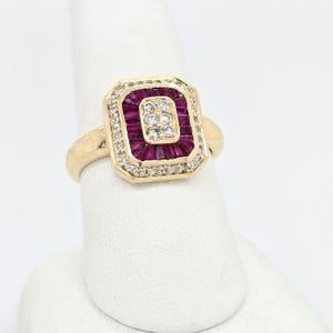 May include: A gold ring with a rectangular face. The face features a central cluster of small diamonds surrounded by a row of ruby baguettes. The outer edge is lined with small diamonds.