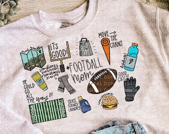 May include: A light grey sweatshirt with a graphic design of football-themed illustrations. The text on the sweatshirt reads "It's good!" "More Cow Bell!" "Football Mom" "He could go all the way!" "Touchdown" "Move the Chains" "Hydrate!" "Good Catch!" "Concessions".