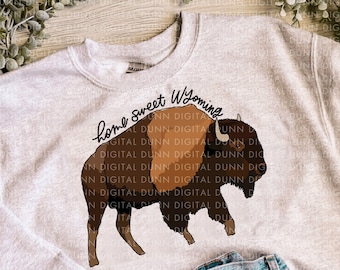 May include: A gray sweatshirt with the text "home sweet Wyoming" and a brown silhouette of a bison. The sweatshirt is laying on a wooden surface with a pair of blue jeans.