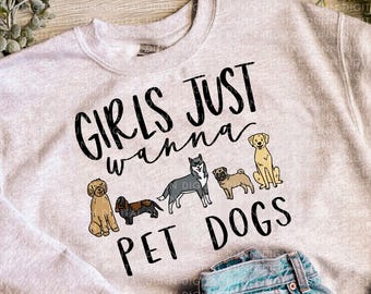 May include: A light gray sweatshirt with the text "GIRLS JUST wanna PET DOGS" in black. The graphic features illustrations of various dog breeds. Light blue denim jeans are folded below the sweatshirt.