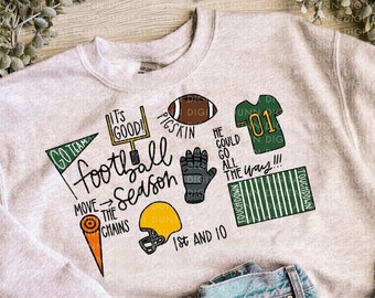 May include: A light gray sweatshirt with colorful football-themed graphics, including a football, helmet, and the words "Football Season." The sweatshirt is paired with light blue denim shorts.