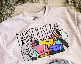 May include: A light gray sweatshirt with the text "Hairstylist life" in black lettering. The design includes a variety of colorful hair styling tools, including scissors, a hair dryer, a comb, and a bottle of shampoo.