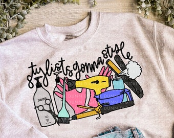May include: A gray sweatshirt with the text "Stylists gonna style" in black lettering. The design includes colorful illustrations of hair styling tools, including scissors, a hair dryer, a comb, and hairspray.