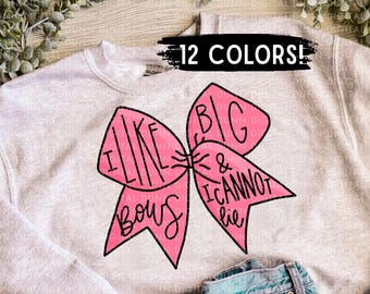 May include: A light gray sweatshirt with a pink bow graphic. The bow has the text "I like big bows & I cannot lie" in black. The text "12 COLORS!" is above the bow. Blue jeans are visible below the sweatshirt.
