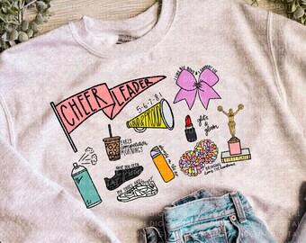 May include: A light gray sweatshirt with a graphic design featuring the text "CHEER LEADER" and other cheerleading-related illustrations. The design includes a megaphone, lipstick, pom-poms, and a trophy. The sweatshirt is paired with blue jeans.
