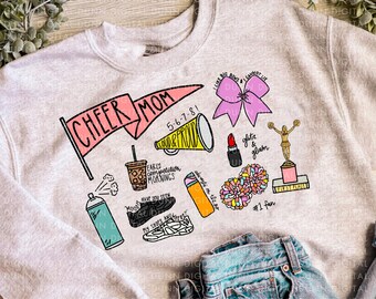 May include: A light gray sweatshirt with various colorful illustrations, including a pink bow, lipstick, and a trophy. The text "CHEER MOM" is displayed on a pink banner. A pair of blue jeans is visible in the image.