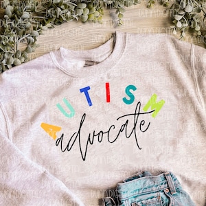 May include: A gray sweatshirt with the words "AUTISM advocate" written in colorful letters. The sweatshirt is laid out on a wooden surface with a pair of brown sandals and a pair of blue jeans.