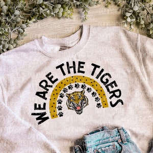 Puede incluir: Una sudadera gris con el texto "We Are The Tigers" en letras negras. El texto está rodeado por un diseño de huellas de patas amarillas y negras.