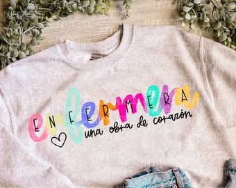 May include: A gray sweatshirt with the text "Enfermera una obra de corazón" in rainbow colors. The text is in a handwritten style with a heart symbol.
