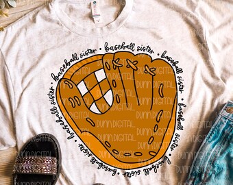 May include: A white t-shirt with a brown baseball glove graphic and the text "baseball sister" repeated around the glove. The shirt is displayed on a wooden surface with a pair of black sandals and blue jeans.
