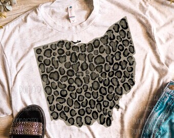 May include: A white t-shirt featuring a grey and black leopard print outline of the state of Ohio. The shirt is lying on a wooden surface with a pair of black sandals with rhinestones.