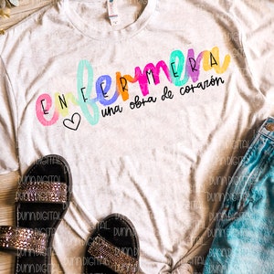 May include: A white t-shirt with the text "ENFERMERA una obra de coraz&oacute;n" in rainbow colors. The text is written in a playful, handwritten style with a heart symbol.