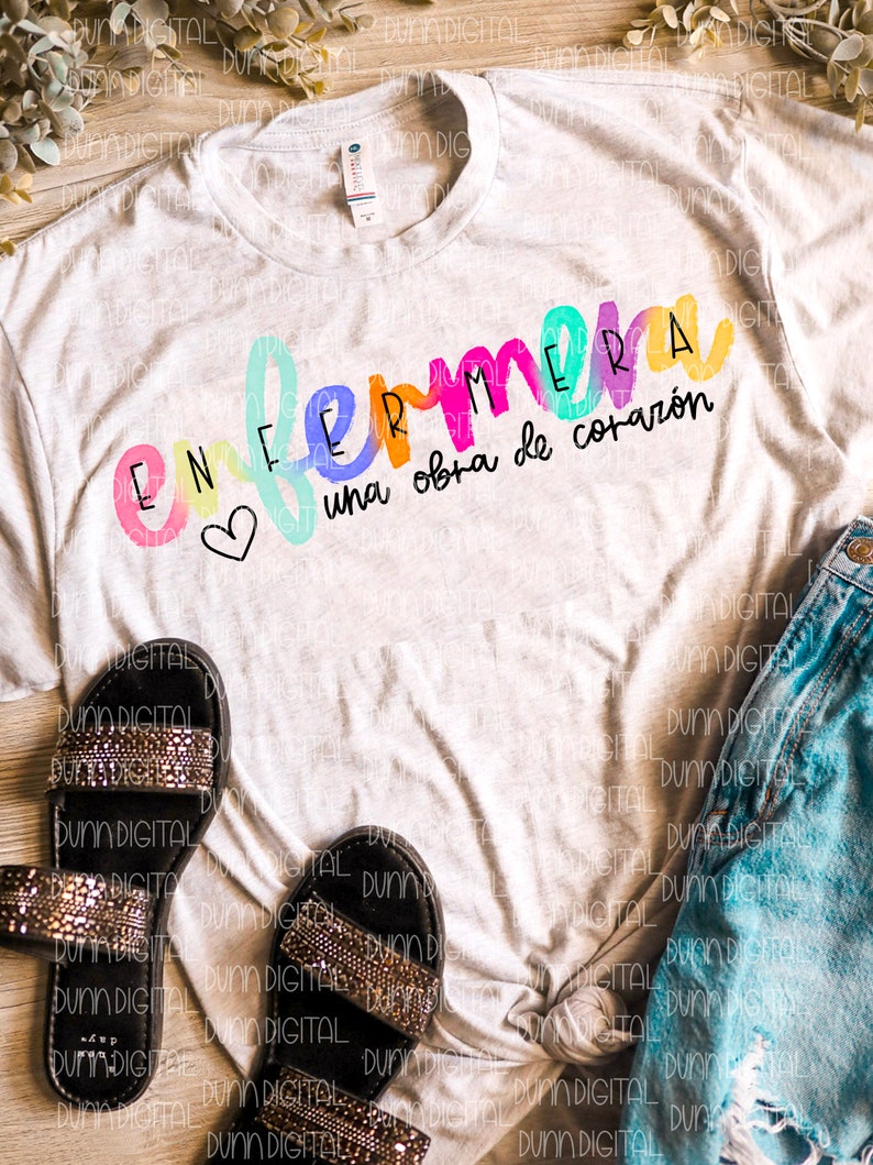 May include: A white t-shirt with the text "ENFERMERA una obra de coraz&oacute;n" in rainbow colors. The text is written in a playful, handwritten style with a heart symbol.