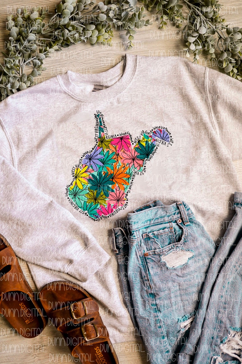 K&ouml;nnte beinhalten: Ein graues Sweatshirt mit einem floralen Design des Bundesstaates West Virginia. Das Design ist mit den Worten "West Virginia" umrandet, die sich um die Form des Bundesstaates wiederholen. Das Sweatshirt liegt auf einer Holzoberfl&auml;che mit einer blauen Jeans und braunen Sandalen.