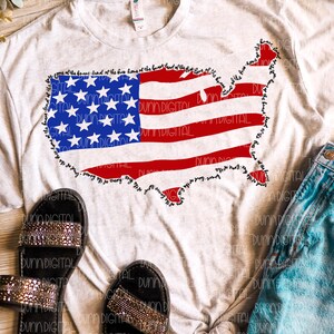 May include: A light gray t-shirt featuring a graphic of the United States in the shape of the American flag. The flag design includes red stripes, blue stars, and the text "land of the free, home of the brave" around the border. Black sandals and denim shorts are also visible.