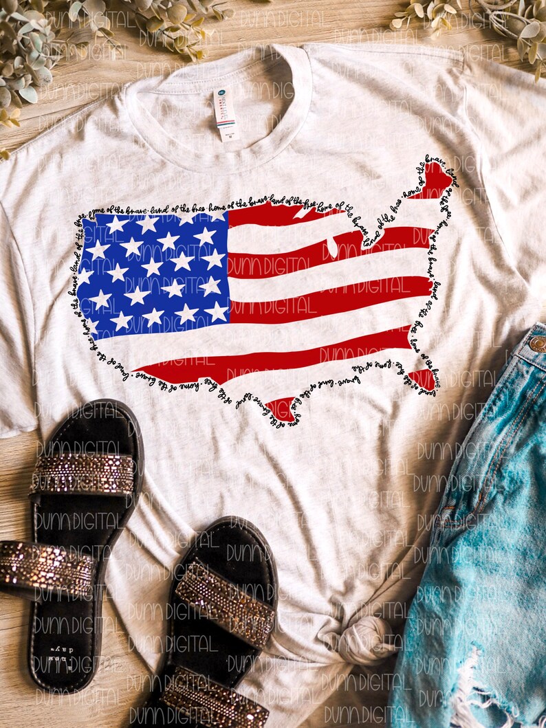 May include: A light gray t-shirt featuring a graphic of the United States in the shape of the American flag. The flag design includes red stripes, blue stars, and the text "land of the free, home of the brave" around the border. Black sandals and denim shorts are also visible.