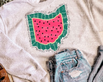 May include: A grey sweatshirt featuring a watermelon-shaped outline of the state of Ohio. The outline is surrounded by the word "Ohio" repeated multiple times. The sweatshirt is laid out on a wooden surface with a pair of blue jeans and brown sandals.