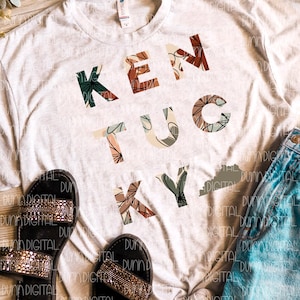 K&ouml;nnte beinhalten: Ein wei&szlig;es T-Shirt mit einem floralen Design, das "Kentucky" buchstabiert, mit einem kleinen Umriss des Bundesstaates Kentucky. Das Shirt liegt auf einer Holzoberfl&auml;che mit einem Paar schwarzer Sandalen und einer blauen Jeans.