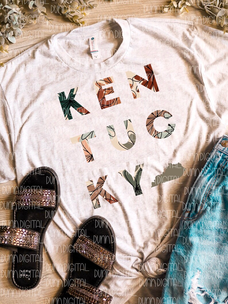 K&ouml;nnte beinhalten: Ein wei&szlig;es T-Shirt mit einem floralen Design, das "Kentucky" buchstabiert, mit einem kleinen Umriss des Bundesstaates Kentucky. Das Shirt liegt auf einer Holzoberfl&auml;che mit einem Paar schwarzer Sandalen und einer blauen Jeans.