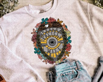 May include: A light gray sweatshirt featuring a floral police badge design. The badge reads "PROUD POLICE WIFE" and is surrounded by colorful flowers. Light blue jeans are visible in the image.