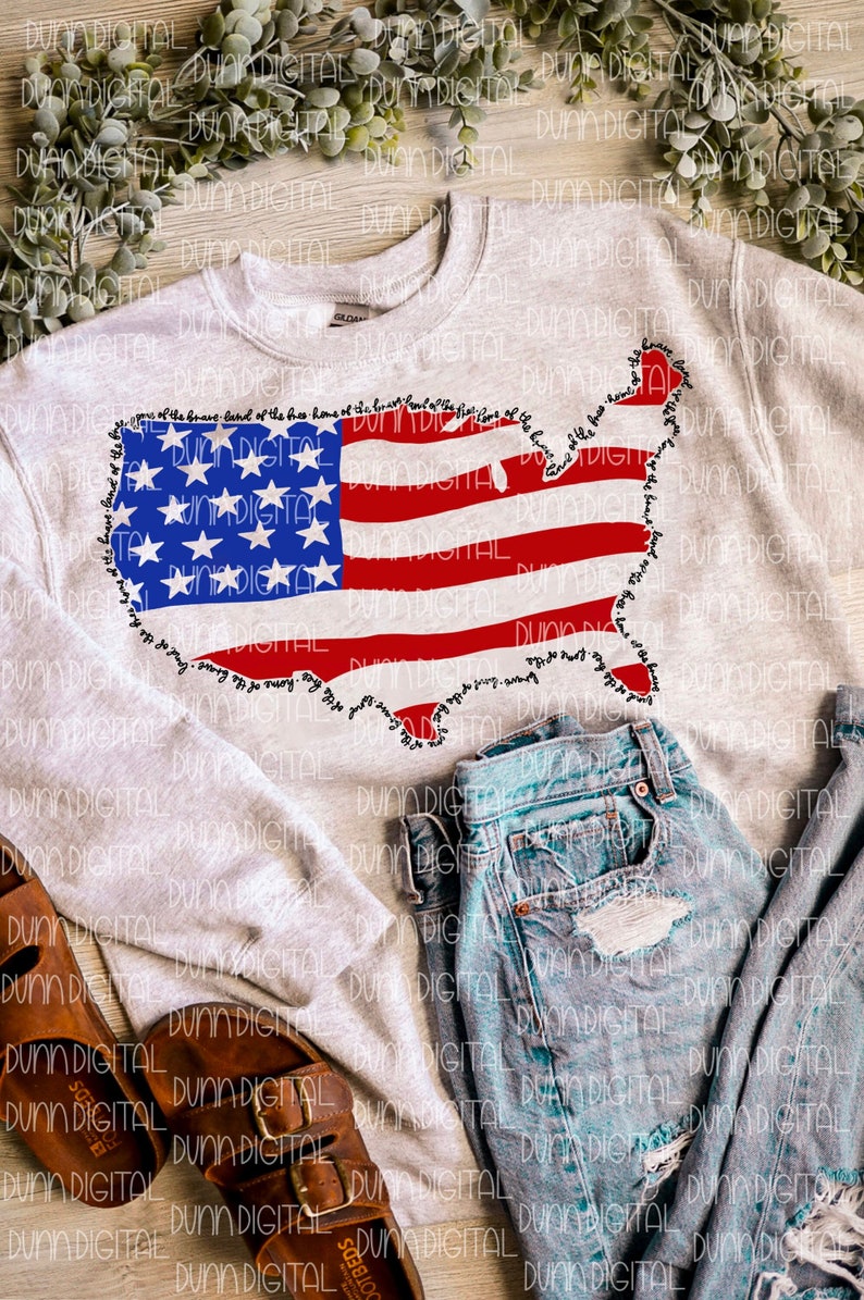 May include: A light gray sweatshirt featuring a graphic of the United States flag in the shape of the country. The flag design includes blue stars and red and white stripes. The words "home of the brave" are printed around the flag's outline. The sweatshirt is paired with blue denim shorts and brown sandals.