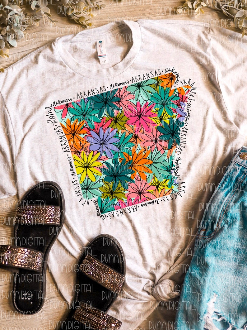 May include: A white t-shirt with a floral design in the shape of the state of Arkansas. The design features a variety of colors, including pink, yellow, orange, blue, and green. The text "Arkansas" is written around the outline of the state.