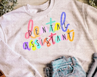 May include: A light gray sweatshirt with the words "DENTAL ASSISTANT" in colorful, handwritten-style lettering. The sweatshirt is paired with light-wash denim jeans and brown sandals.