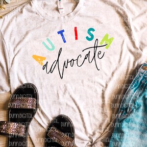 May include: A white t-shirt with the words "AUTISM advocate" in colorful letters. The letters are in different colors, including blue, red, green, yellow, and orange. The words are written in a cursive font.