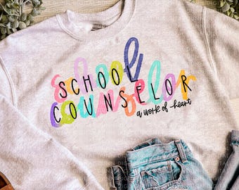 May include: A light gray sweatshirt with the words "SCHOOL COUNSELOR" in colorful letters and "a work of heart" in black. The sweatshirt is paired with light blue denim jeans and brown sandals.